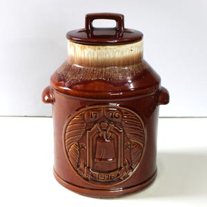 May include: A brown ceramic jar with a lid and handle, featuring a decorative emblem with the numbers "1776" and "1976". The jar has a milk can shape with a light brown and white top.