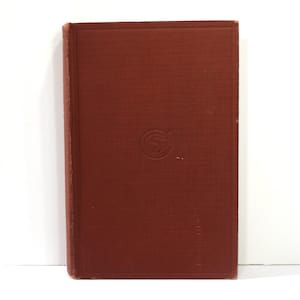 May include: A vintage book with a textured, reddish-brown cover. The cover features a debossed circular design in the center. The book's spine is visible, and the edges show signs of wear, suggesting its age. The background is plain white.