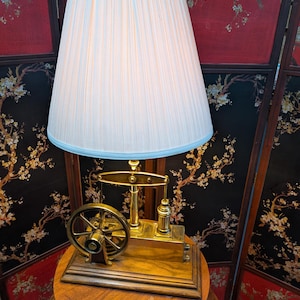 May include: A vintage-style table lamp with a cream-colored pleated lampshade. The lamp features a brass-colored, industrial-inspired base with a flywheel and other mechanical details, mounted on a wooden base. The lamp is placed on a round wooden table.