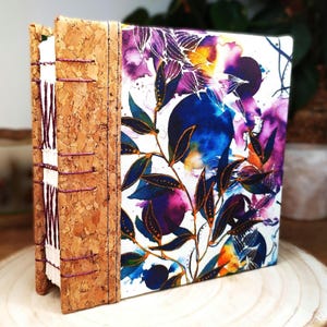 May include: A handmade book with a cork spine and exposed purple thread stitching. The front cover features a vibrant, abstract botanical print with deep blue, purple, and orange watercolor-style foliage patterns on a white background.