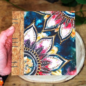 May include: A square journal with a cork spine and a colorful floral design cover. The cover features a mandala-style pattern with shades of blue, red, yellow, and white. The journal is held in a person's hand.