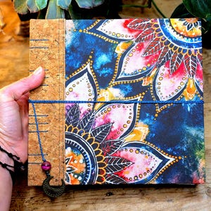 May include: A handmade journal with a cork spine and a vibrant floral design on the cover. The cover features a blue background with colorful mandala-style flowers in shades of pink, yellow, and white. A blue cord wraps around the journal, with a charm.