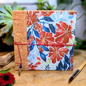May include: A square journal with a cork spine and a floral fabric cover featuring red and blue flowers on a light blue background. A red cord wraps around the journal, and a feather charm hangs from the spine. Paintbrushes rest on a wooden surface.