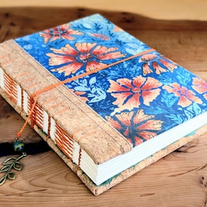 May include: A hardcover journal featuring a blue fabric cover with an orange floral pattern. The spine and edges are made of textured cork material, bound with visible orange thread stitching. An orange cord bookmark with a metal leaf charm is attached.