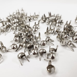 May include: A large quantity of silver metal double-pronged rivets. The rivets have a round head and are scattered on a white surface.