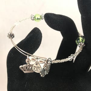 Puede incluir: Un brazalete de plata con cuentas verdes y transparentes. El brazalete tiene un dije que dice "hecho con amor" y un dije de corona.