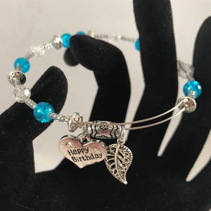 Puede incluir: Un brazalete de plata con cuentas azules y transparentes. El brazalete tiene un dije en forma de corazón que dice "Feliz cumpleaños" y un dije en forma de hoja.