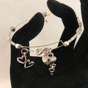 Puede incluir: Un brazalete de plata con un dije de símbolo de infinito, un dije de corazón con un ojo de cerradura y dos dijes de corazón más pequeños. El brazalete está hecho de una cadena de plata con pequeñas cuentas.