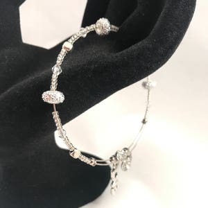 Puede incluir: Una pulsera plateada con cuentas y adornos claros y brillantes. La pulsera presenta un diseño delicado con múltiples cuentas pequeñas, redondas y ovaladas. La pulsera se muestra sobre un fondo negro.