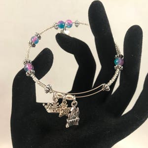 Puede incluir: Un brazalete extensible de plata con cuentas iridiscentes de color rosa y azul y abalorios. Los abalorios incluyen una corona y una pequeña casa con la palabra "Home" en ella.