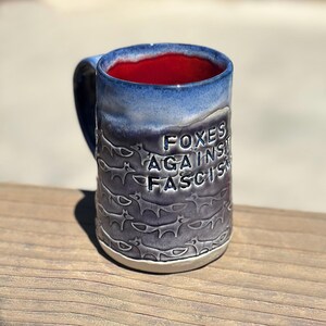 May include: A ceramic mug with a blue and red interior. The exterior features a repeating fox pattern and the words "FOXES AGAINST FASCISM" in embossed lettering. The mug has a dark blue handle and a white base.