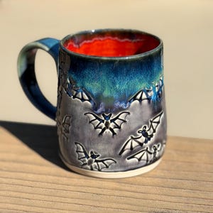 May include: A ceramic mug featuring a relief pattern of flying bats across a grey body. The upper portion of the mug transitions into a deep blue glaze, while the interior is finished in a vibrant, solid orange colour with a dark blue rim.
