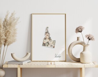 May include: A framed print of the state of Idaho with a floral design in shades of beige, brown, and green. The print is on a white background and the word "Idaho" is written in cursive at the bottom. The print is displayed on a light wood shelf with two decorative vases and dried pampas grass.