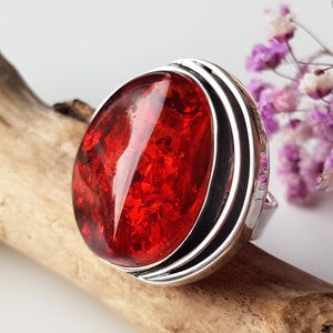 May include: A ring featuring a large, oval-shaped, deep red translucent stone with internal inclusions, set within a polished silver-toned metal band that has multiple layered grooves along the side of the setting.