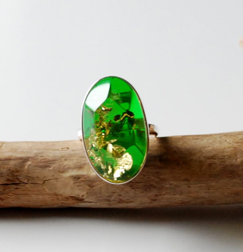 K&ouml;nnte beinhalten: Ein silberner Ring mit einem gro&szlig;en ovalen gr&uuml;nen Edelstein mit goldenen Flocken im Inneren. Der Ring liegt auf einem St&uuml;ck Treibholz.