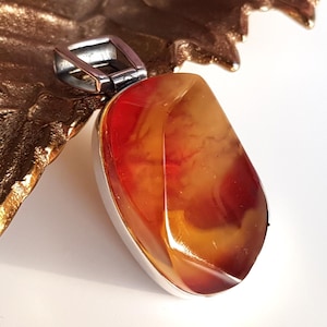 Puede incluir: Un colgante con una piedra grande, pulida, de forma ovalada en tonos rojos, naranjas y amarillos. La piedra está engastada en un marco de metal plateado con una fianza rectangular. El colgante se exhibe sobre una superficie dorada texturizada.