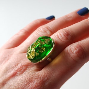 K&ouml;nnte beinhalten: Ein silberner Ring mit einem gro&szlig;en ovalen gr&uuml;nen Stein, der darin eingefasst ist. Der Stein hat goldene Flecken.
