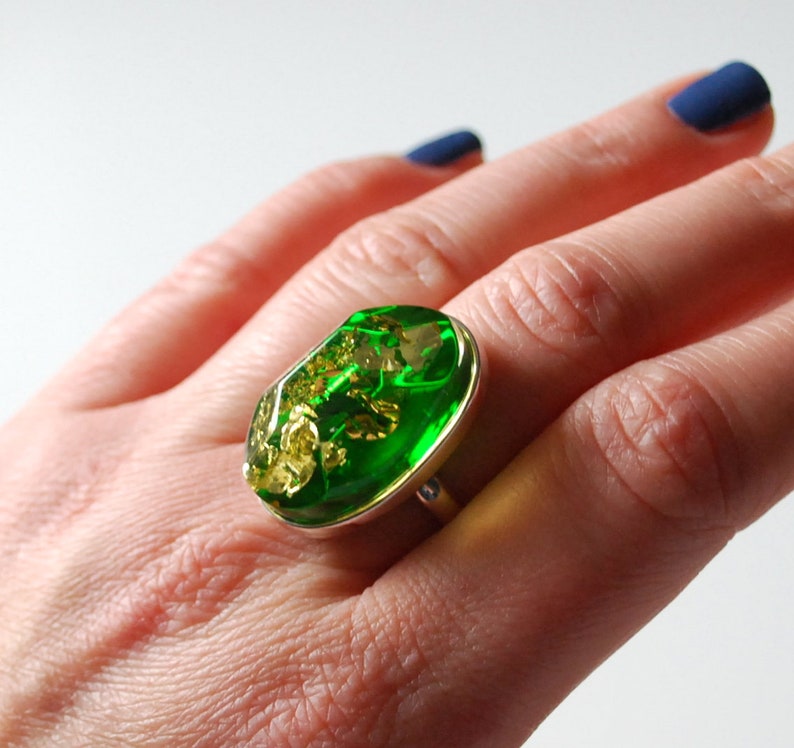 K&ouml;nnte beinhalten: Ein silberner Ring mit einem gro&szlig;en ovalen gr&uuml;nen Stein, der darin eingefasst ist. Der Stein hat goldene Flecken.