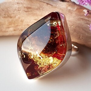 Könnte beinhalten: Ein auffälliger Ring mit einem großen, facettierten, rotbraunen Stein mit Goldeinlagerungen. Der Stein ist in einem silberfarbenen Band gefasst, was einen auffälligen Kontrast erzeugt. Das Design des Rings ist modern und auffällig.