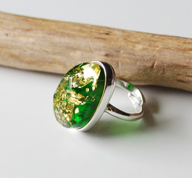 K&ouml;nnte beinhalten: Ein silberner Ring mit einem ovalen, gr&uuml;n-goldenen Stein. Der Stein ist aus Harz und hat goldene Flocken eingebettet.