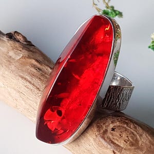 Op de afbeelding: Een ring met een grote, langwerpige, doorschijnende rode steen in een gepolijste zilveren zetting. De ringband heeft een getextureerde, schorsachtige afwerking die contrasteert met het gladde, levendige rode oppervlak van de edelsteen.