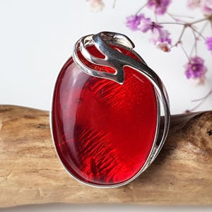 May include: Oval-shaped red gemstone pendant with a silver-colored metal frame. The pendant has a textured, internal pattern. The silver frame curves around the gemstone. The pendant is displayed on a piece of wood.