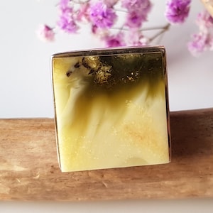 Könnte beinhalten: Ein quadratisches Stück Seife mit einem geschichteten Design in Weiß, Gelb und Grün. Die Seife hat einen goldenen Rand und liegt auf Holz. Im Hintergrund sind lila Blumen zu sehen. Handgemachte Körperpflege.