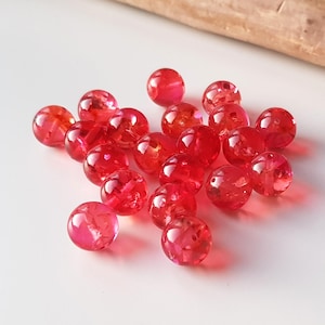 Puede incluir: Un conjunto de cuentas rojas translúcidas, cada una de aproximadamente 1,3 cm de diámetro, dispersas sobre una superficie blanca. Las cuentas tienen un acabado brillante y parecen estar hechas de vidrio o un material similar. Un trozo de madera está en el fondo.