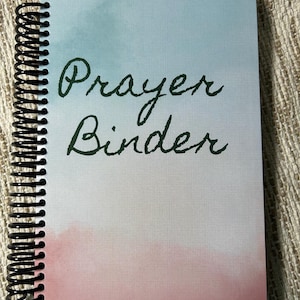 May include: A spiral-bound prayer binder with a watercolor design. The cover features a gradient of pink and blue hues, with the words "Prayer Binder" in dark green script. The binder's spine is black.
