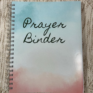 May include: A spiral-bound notebook with a watercolor design. The cover features a gradient of blue, white, and pink hues. The words "Prayer Binder" are written in dark green script across the front.