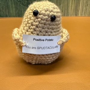 May include: A beige crocheted potato figure with two small black bead eyes. The figure holds a white rectangular sign with black text that reads: Positive Potato. You are SPUDTACULAR! The crocheted object has a rounded, textured surface.