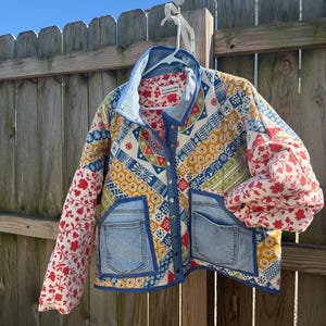 May include: A quilted jacket featuring a patchwork design of red floral, yellow geometric, and blue patterned fabric. The jacket includes light blue denim pockets, a denim collar, and a front snap button closure. A white label is visible inside the collar.