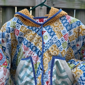 May include: A quilted jacket featuring a vibrant patchwork pattern of blue, mustard yellow, red, and green geometric and floral motifs. The jacket includes a mustard yellow waffle-textured hood, a front button closure, and two large front pockets.