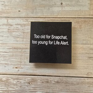 May include: A square black sign with white text that reads: Too old for Snapchat, too young for Life Alert. The sign has a smooth, matte finish and clean, sharp edges, presenting a clear and legible message against the dark background.