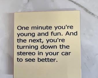 May include: A square, cream-colored coaster featuring black text that reads: One minute you're young and fun. And the next, you're turning down the stereo in your car to see better. The coaster has a smooth, glossy finish and clean, straight edges.