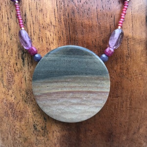 May include: A necklace with a large, round, brown and white stone pendant. The pendant has a layered, striped pattern. The necklace is made of red beads and purple beads.