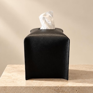 May include: A rectangular black tissue box cover with a smooth, leather-like texture. The cover features a centered top opening through which a single white tissue is pulled upward. The structure has clean, straight edges and a solid black finish.