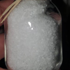 May include: A small glass jar with a cork lid and a straw-like ribbon tied around the top. The jar is filled with white salt crystals.
