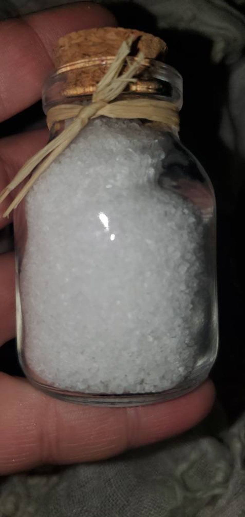 May include: A small glass jar with a cork lid and a straw-like ribbon tied around the top. The jar is filled with white salt crystals.