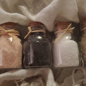 May include: Five small glass jars with cork lids filled with different types of salt. The jars are arranged in a row on a white fabric background.