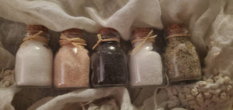 May include: Five small glass jars with cork lids filled with different types of salt. The jars are arranged in a row on a white fabric background.