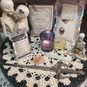 May include: A black and white image of a table setting with a black salt, a small bottle of salt, a candle, a small sword, and two skeletons. The table is covered with a white crocheted tablecloth. The text on the salt packages reads "Black Salt" and "Salt Mean".