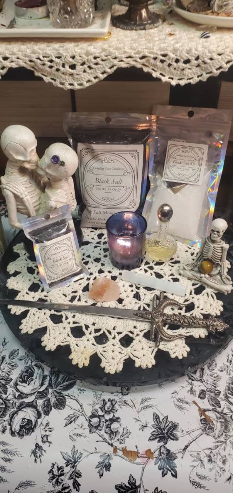 May include: A black and white image of a table setting with a black salt, a small bottle of salt, a candle, a small sword, and two skeletons. The table is covered with a white crocheted tablecloth. The text on the salt packages reads "Black Salt" and "Salt Mean".