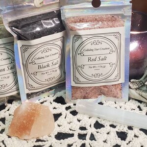 May include: Three small bags of salt labeled "Protection Salt", "Black Salt", and "Red Salt". The bags are on a white crocheted surface with a pink Himalayan salt rock and a white crystal wand.