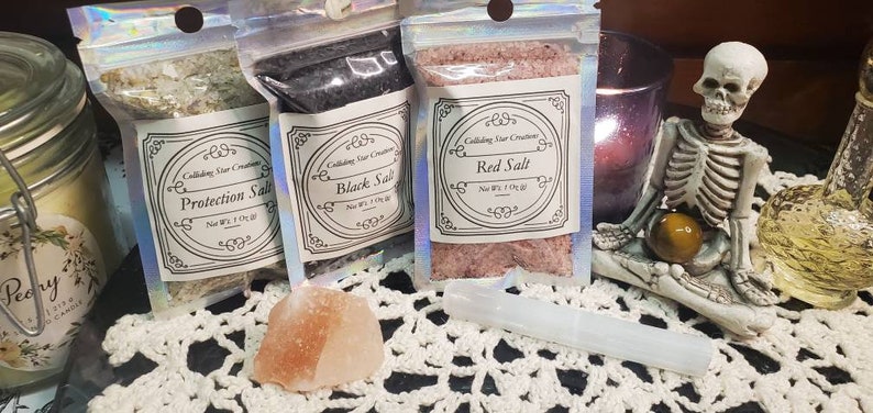 May include: Three small bags of salt labeled "Protection Salt", "Black Salt", and "Red Salt". The bags are on a white crocheted surface with a pink Himalayan salt rock and a white crystal wand.