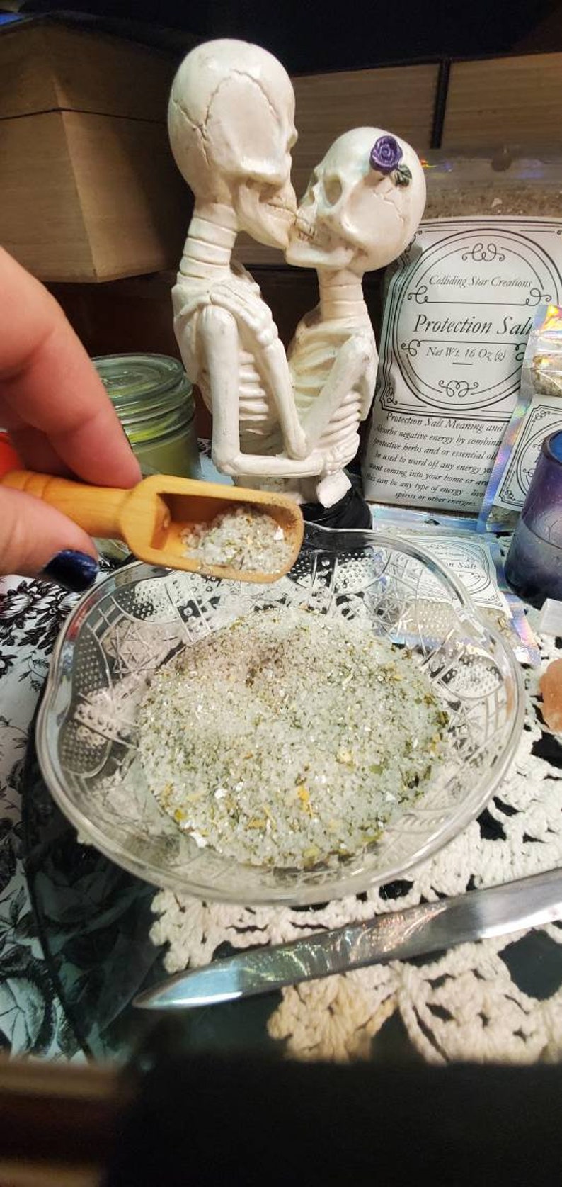 May include: A white ceramic figurine of two skeletons kissing, a glass bowl filled with white salt crystals, and a bag of protection salt with the text "Protection Salt" and "Net Wt. 16 Oz (453g)".