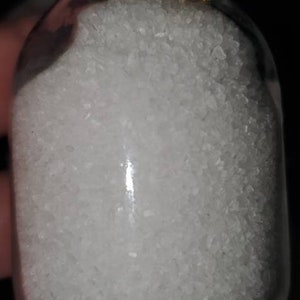 May include: A small glass jar with a cork lid and a brown string tied around the neck. The jar is filled with white crystals.