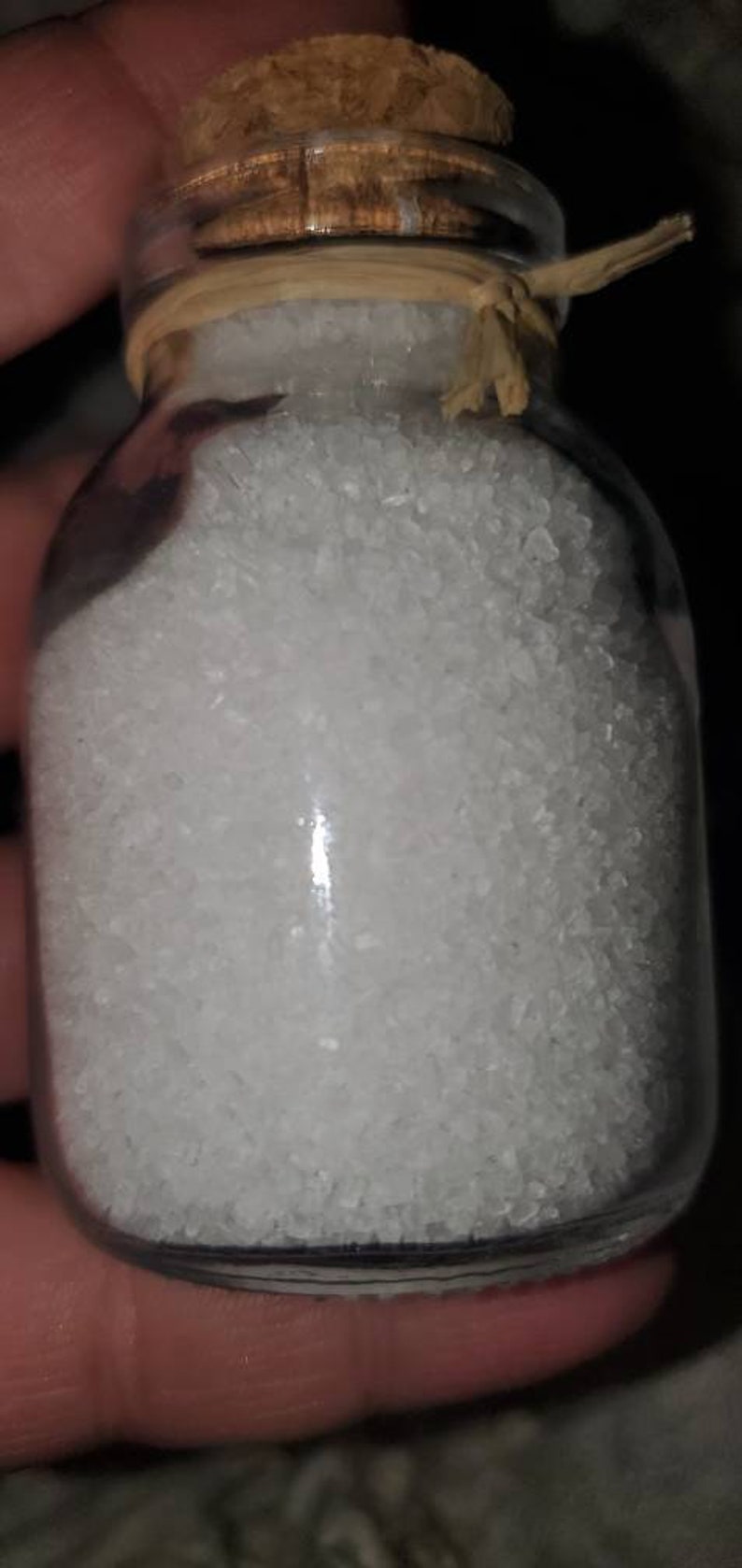 May include: A small glass jar with a cork lid and a brown string tied around the neck. The jar is filled with white crystals.