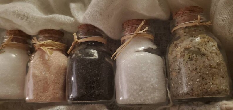 May include: Five small glass jars with cork lids and twine tied around the necks. Each jar contains a different type of salt: white, pink, black, and two different shades of brown.