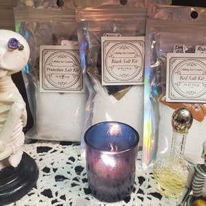 May include: Three clear plastic bags containing salt kits for various purposes. The bags are labeled "Protection Salt Kit", "Black Salt Kit", and "Red Salt Kit". The kits are displayed on a white lace tablecloth with a small glass jar of green liquid, a small glass bottle with a gold top, and a small glass candle holder with a purple candle.